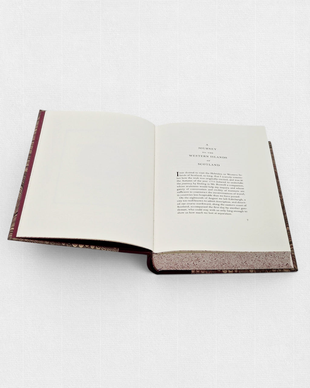 Otwarta książka „A Journey to the Western Islands of Scotland” z widoczną stroną tytułową rozdziału i pierwszym akapitem tekstu; po lewej stronie pusta karta, na dole widoczne jasne, nakrapiane brzegi stron oraz fragment marmurkowej oprawy.