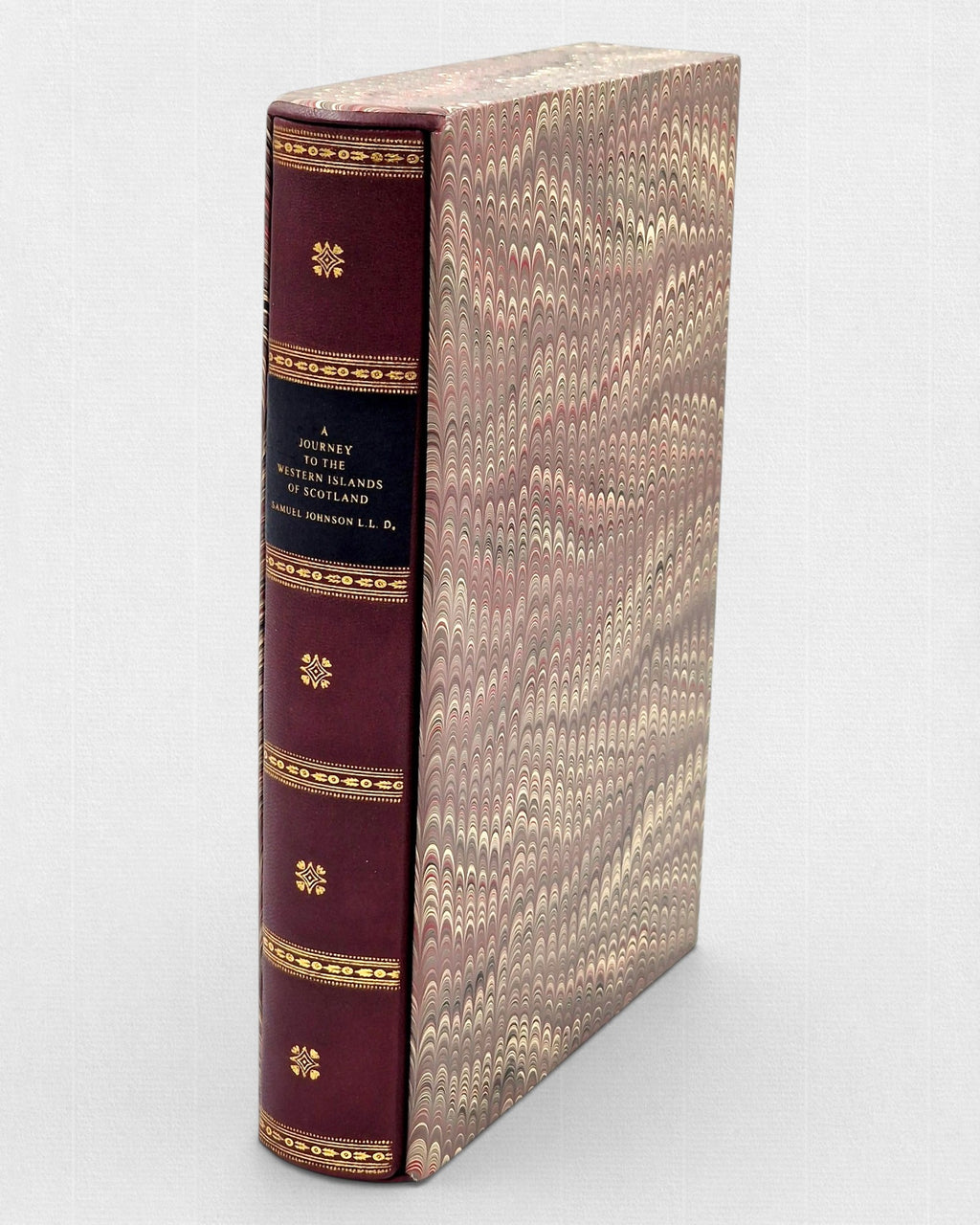 Bordowa, skórzana książka „A Journey to the Western Islands of Scotland” ze złoconymi zdobieniami na grzbiecie, wsunięta w ozdobne etui pokryte papierem marmurkowym w odcieniach brązu, beżu i czerwieni.