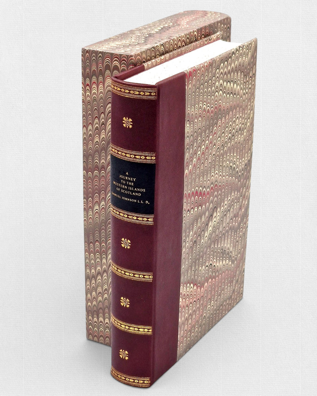 Bordowa, skórzana książka „A Journey to the Western Islands of Scotland” ze złoconymi zdobieniami na grzbiecie, częściowo wysunięta z ozdobnego etui pokrytego papierem marmurkowym w odcieniach brązu, beżu i czerwieni.