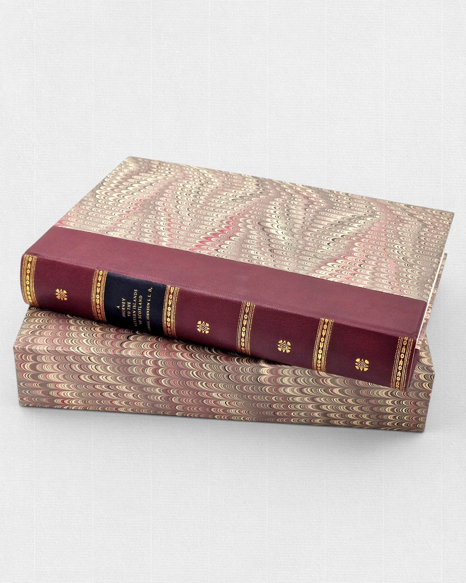 Bordowa, skórzana książka „A Journey to the Western Islands of Scotland” ze złoconymi zdobieniami na grzbiecie, ułożona poziomo na dopasowanym etui pokrytym papierem marmurkowym w odcieniach beżu, brązu i czerwieni.