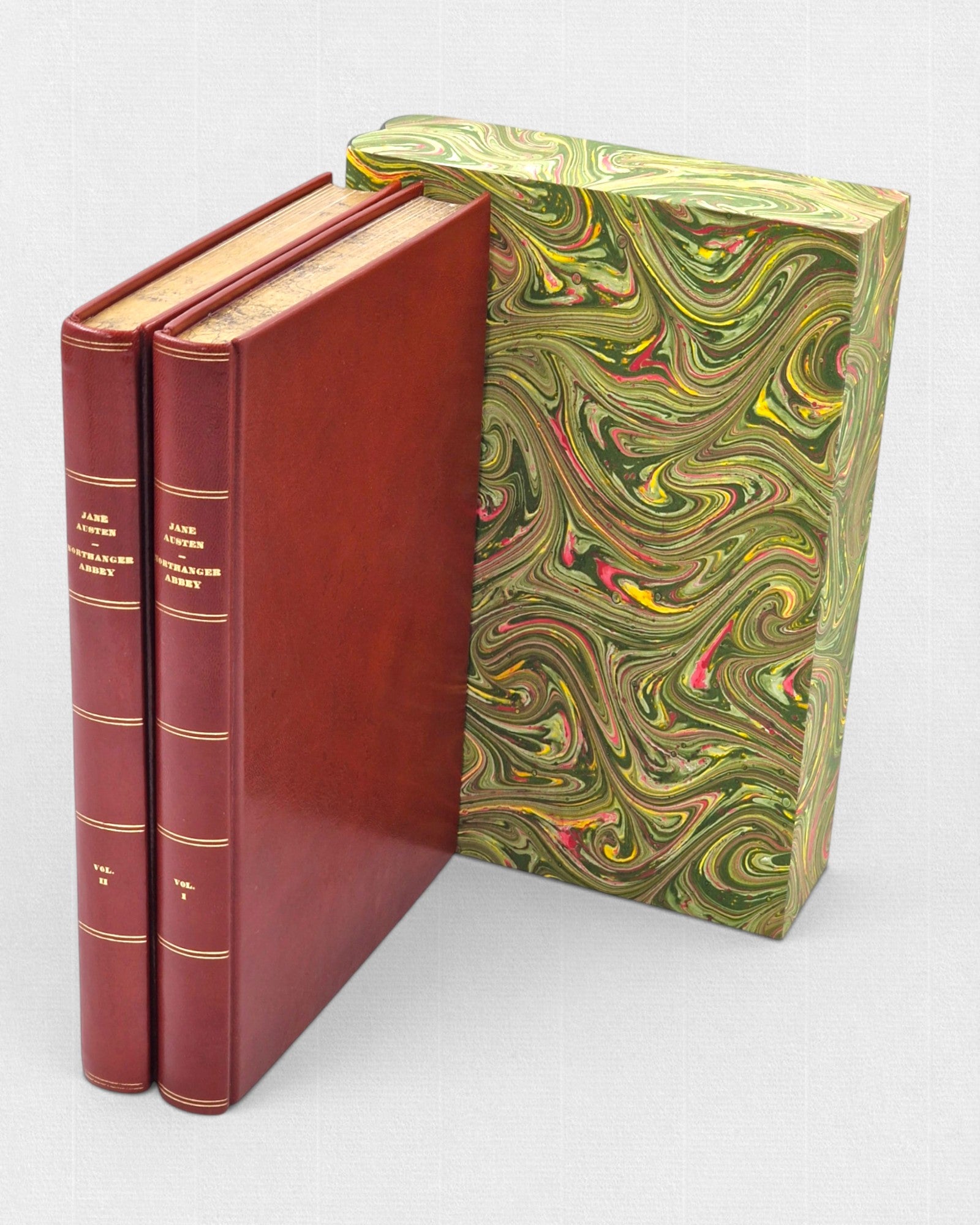 Dwutomowe wydanie „Northanger Abbey” Jane Austen w czerwonej skórzanej oprawie ze złoceniami, obok dekoracyjnego etui w marmurkowy wzór w odcieniach zieleni, żółci i czerwieni.