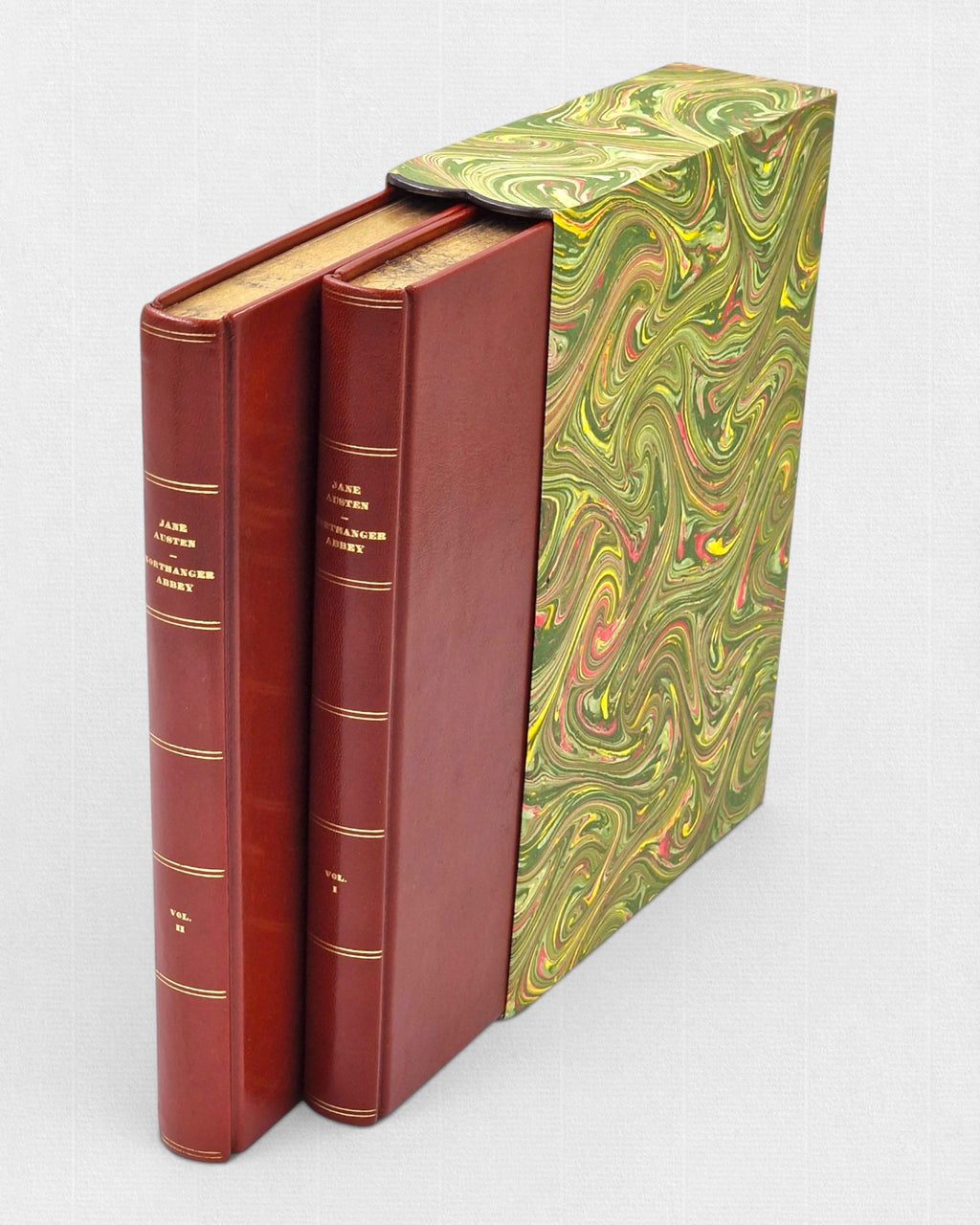 Dwutomowe wydanie „Northanger Abbey” Jane Austen w czerwonej skórzanej oprawie ze złoconymi napisami na grzbietach, częściowo wsunięte w etui w marmurkowy wzór w odcieniach zieleni, żółci i czerwieni.